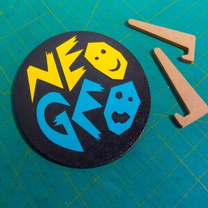 Może przedstawiać: Czarny okrągły dysk z napisem "NEO GEO" w ż&oacute;łtych i niebieskich literach. Litery są obwiedzione białym kolorem i mają fakturowaną, błyszczącą powierzchnię. Na dysku znajdują się dwie ż&oacute;łte animowane twarze.