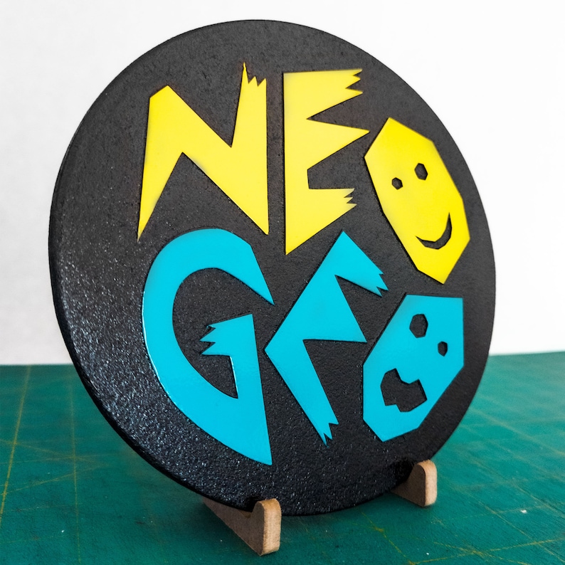 Może przedstawiać: Okrągły czarny znak z napisem "NEO GEO" w ż&oacute;łtych i niebieskich literach. Litery są stylizowane i mają ząbkowane krawędzie. Znak stoi na drewnianym stojaku.