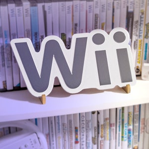 Puede incluir: Un letrero blanco con la palabra "Wii" en letras grises, de pie en un estante con filas de cajas de videojuegos detrás.