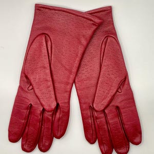 Puede incluir: Un par de guantes de cuero rojo con un dise&ntilde;o perforado. Los guantes son de un tono rojo vibrante y parecen estar hechos de cuero suave. Los guantes est&aacute;n planos sobre una superficie blanca.