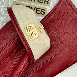 Puede incluir: Un par de guantes de cuero rojo con un dise&ntilde;o perforado. Los guantes est&aacute;n forrados con forro polar de algod&oacute;n y tienen una etiqueta que dice "Real Leather Gloves Lined Cotton Fleece Made in England". Tambi&eacute;n es visible una etiqueta de talla 7.