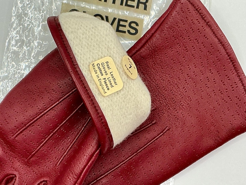 Puede incluir: Un par de guantes de cuero rojo con un dise&ntilde;o perforado. Los guantes est&aacute;n forrados con forro polar de algod&oacute;n y tienen una etiqueta que dice "Real Leather Gloves Lined Cotton Fleece Made in England". Tambi&eacute;n es visible una etiqueta de talla 7.