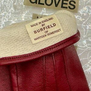 Puede incluir: Primer plano de guantes de cuero rojo con pu&ntilde;o blanco. Los guantes tienen una superficie texturizada y una etiqueta que dice "Made in England by Burfield of Martock Somerset."