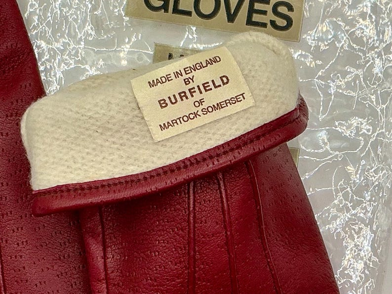 Puede incluir: Primer plano de guantes de cuero rojo con pu&ntilde;o blanco. Los guantes tienen una superficie texturizada y una etiqueta que dice "Made in England by Burfield of Martock Somerset."