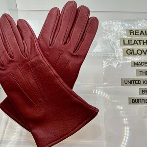 Puede incluir: Un par de guantes de cuero rojo con peque&ntilde;as perforaciones. Los guantes se exhiben sobre una superficie transparente. El texto en el embalaje dice "REAL LEATHER GLOVES MADE IN THE UNITED KINGDOM BY BURFIELD'S."