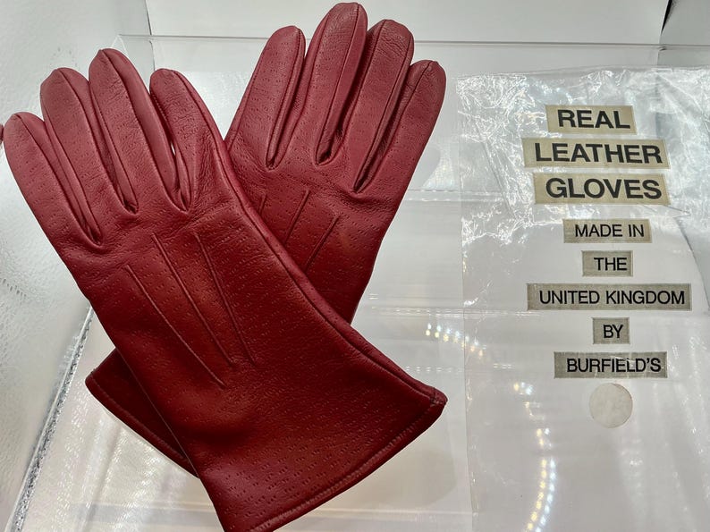 Puede incluir: Un par de guantes de cuero rojo con peque&ntilde;as perforaciones. Los guantes se exhiben sobre una superficie transparente. El texto en el embalaje dice "REAL LEATHER GLOVES MADE IN THE UNITED KINGDOM BY BURFIELD'S."