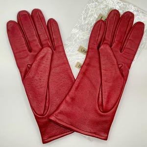 Puede incluir: Un par de guantes de cuero rojo brillante. Los guantes tienen un dise&ntilde;o perforado en la parte posterior y est&aacute;n sobre una superficie blanca. Los guantes son nuevos y est&aacute;n en su embalaje original.