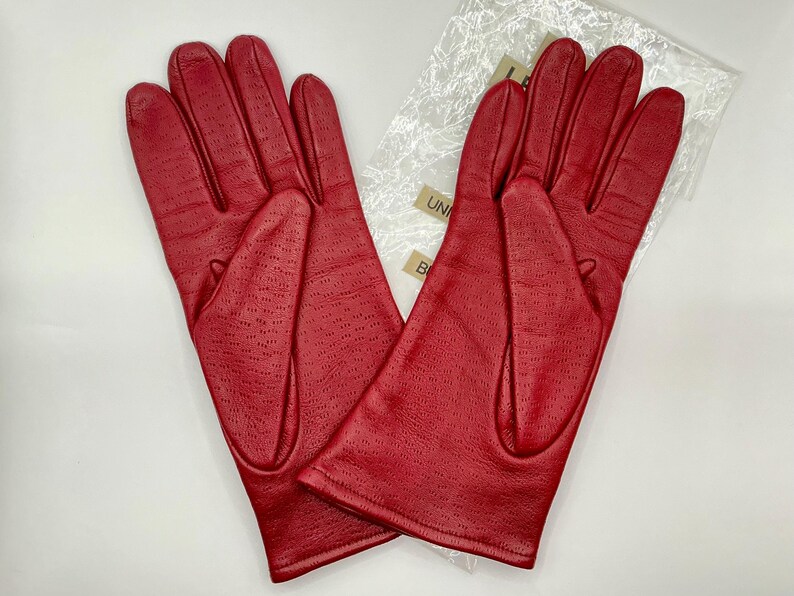 Puede incluir: Un par de guantes de cuero rojo brillante. Los guantes tienen un dise&ntilde;o perforado en la parte posterior y est&aacute;n sobre una superficie blanca. Los guantes son nuevos y est&aacute;n en su embalaje original.