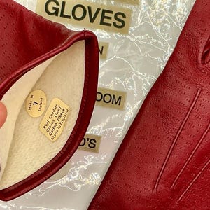 Puede incluir: Primer plano de un par de guantes de cuero rojo. Los guantes est&aacute;n perforados y tienen un forro de suave forro polar de algod&oacute;n. La etiqueta dentro del guante dice "Real Leather Gloves Lined Cotton Fleece Made in England".