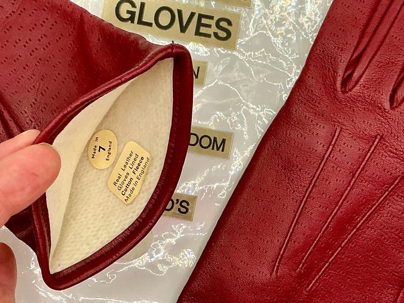 Puede incluir: Primer plano de un par de guantes de cuero rojo. Los guantes est&aacute;n perforados y tienen un forro de suave forro polar de algod&oacute;n. La etiqueta dentro del guante dice "Real Leather Gloves Lined Cotton Fleece Made in England".