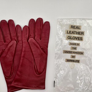 Puede incluir: Un par de guantes de cuero burdeos, junto a una bolsa de pl&aacute;stico transparente con el texto "REAL LEATHER GLOVES MADE IN THE UNITED KINGDOM BY BURFIELD'S". Los guantes son nuevos.