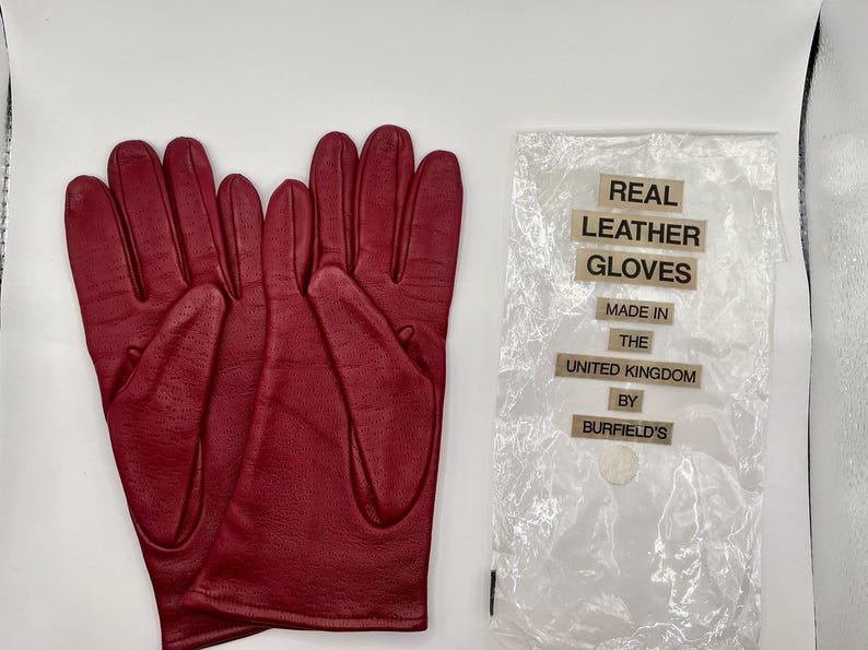 Puede incluir: Un par de guantes de cuero burdeos, junto a una bolsa de pl&aacute;stico transparente con el texto "REAL LEATHER GLOVES MADE IN THE UNITED KINGDOM BY BURFIELD'S". Los guantes son nuevos.