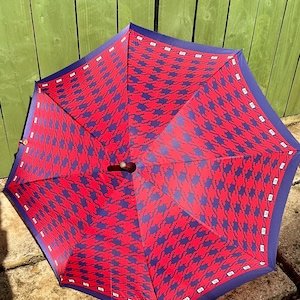 May include: An open umbrella featuring a vibrant red and dark blue houndstooth pattern across its canopy. The edges are finished with a solid dark blue border containing a repeating white square motif. A dark wooden handle is visible at the center.