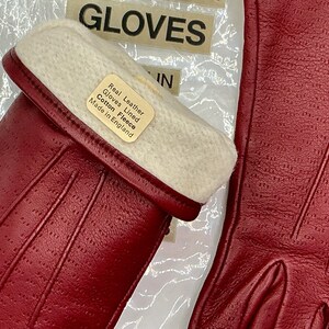 Puede incluir: Primer plano de guantes de cuero rojo con forro de forro polar blanco. Los guantes tienen una superficie texturizada y est&aacute;n etiquetados como "Real Leather Gloves Lined Cotton Fleece Made in England". La palabra "GLOVES" es visible en el fondo.