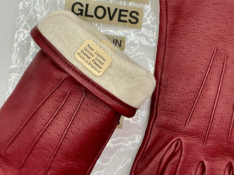 Puede incluir: Primer plano de guantes de cuero rojo con forro de forro polar blanco. Los guantes tienen una superficie texturizada y est&aacute;n etiquetados como "Real Leather Gloves Lined Cotton Fleece Made in England". La palabra "GLOVES" es visible en el fondo.