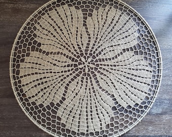 May include: A circular beige crochet doily featuring a central floral motif surrounded by intricate radiating fan-shaped patterns and a delicate hexagonal mesh border, all mounted on a thin, matching circular frame for structural support.
