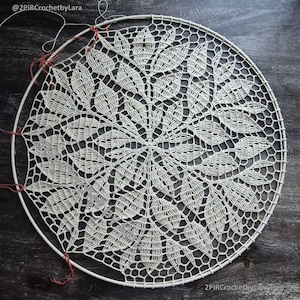 May include: Cream-colored, handmade crochet doily with a leaf pattern, stretched over a white metal hoop. The intricate design features a central floral motif surrounded by detailed leaf shapes. The piece is partially complete, with loose threads visible.