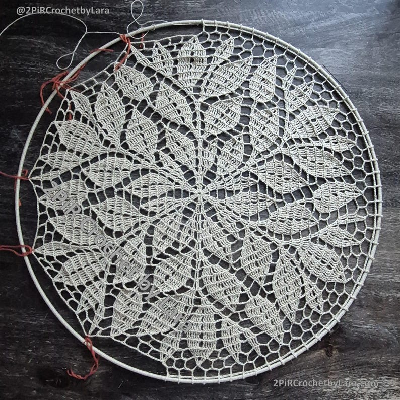 May include: Cream-colored, handmade crochet doily with a leaf pattern, stretched over a white metal hoop. The intricate design features a central floral motif surrounded by detailed leaf shapes. The piece is partially complete, with loose threads visible.
