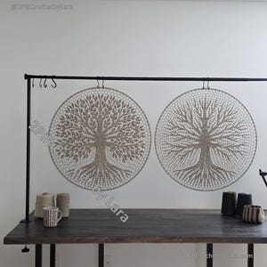 Puede incluir: Dos tapices de pared circulares con diseños de árboles. Los árboles están intrincadamente tejidos a ganchillo en un color beige neutro, con ramas y raíces detalladas. Los tapices están suspendidos de una barra de metal negro, creando una estética natural y terrosa.