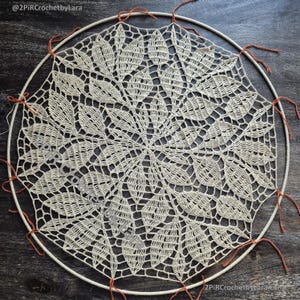 May include: Cream-colored, handmade crochet doily with a floral design, stretched over a circular wooden hoop. The doily is secured with orange thread. The text "@2PIRCrochetbyLara" is visible in the image.