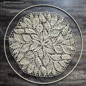 May include: A cream-colored, handmade crochet doily with a detailed floral pattern, set within a white embroidery hoop. The doily's intricate design is centered, showcasing the delicate craftsmanship. The image is set against a dark wood grain background.