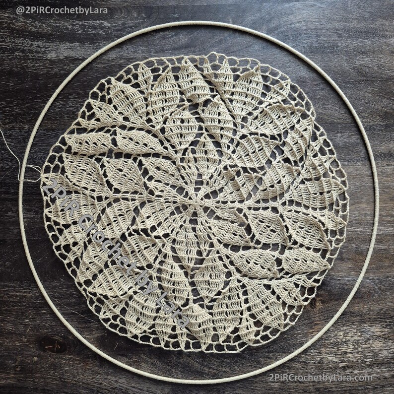 May include: A cream-colored, handmade crochet doily with a detailed floral pattern, set within a white embroidery hoop. The doily's intricate design is centered, showcasing the delicate craftsmanship. The image is set against a dark wood grain background.