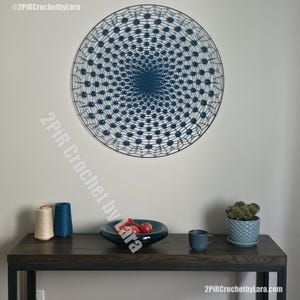 Op de afbeelding: Een grote ronde wanddecoratie met een donkerblauw gehaakt mandalapatroon gespannen in een dunne zwarte metalen ring. Het ingewikkelde ontwerp bestaat uit herhalende radiale motieven en webachtige structuren voor een symmetrisch geometrisch geheel.