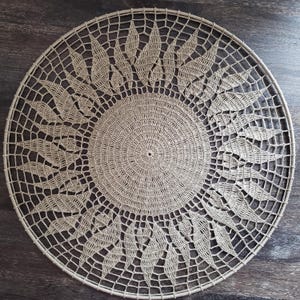 Pode incluir: Um tapete de crochê circular bege com um centro sólido e de ponto apertado, rodeado por um padrão intrincado e aberto de formas radiantes semelhantes a folhas. A peça está montada numa moldura circular a condizer, criando uma decoração de parede estruturada, plana e simétrica com um design de fibra detalhado e repetitivo.