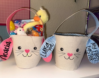 Puede incluir: Dos cestas de Pascua con caras de conejito. Una cesta es rosa y dice "Acacia" y la otra es azul y dice "Mi primera Pascua".