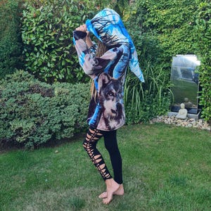 May include: A long-sleeved hooded sweatshirt with a wolf graphic on the front. The sweatshirt has a blue and white design on the hood and sleeves. The person is wearing black leggings with cut-out details. The background is a green lawn and foliage.