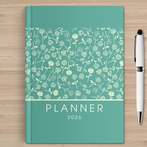 May include: A teal planner with a floral pattern and the word "PLANNER" in white. The year "2026" is below the word. A white pen with silver accents rests on a wooden surface next to the planner.