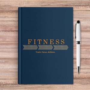 May include: A dark blue notebook with the word "FITNESS" in orange text and the words "Track. Focus. Achieve." in smaller orange text. The notebook is on a wooden surface with a silver pen next to it.