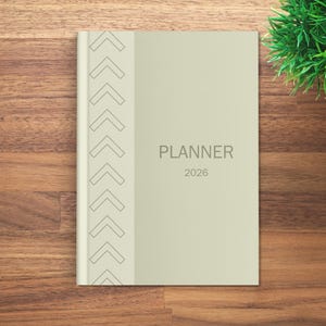 May include: A light green 2026 planner with the word "PLANNER" printed in gray. The planner has a decorative arrow design on the left side. The planner is on a wooden surface.