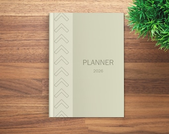 May include: A light green 2026 planner with the word "PLANNER" printed in gray. The planner has a decorative arrow design on the left side. The planner is on a wooden surface.