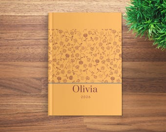 May include: A mustard-colored journal with a floral pattern and the name "Olivia" and the year "2026" printed on the cover. The floral design features brown roses and stems. A thin gray line separates the floral pattern from the name and year.