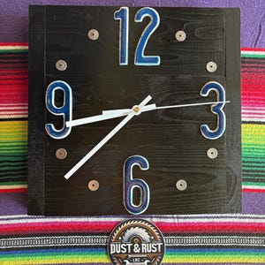 May include: Square black wooden clock with blue numbers and white hands. The numbers 3, 6, 9, and 12 are visible. The clock has a sticker that says "Dust & Rust - LBC". The clock is on a colorful striped background.