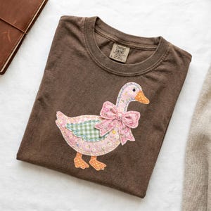 Peut inclure: T-shirt Comfort Colors marron avec une application décorative d'oie. L'oie a un corps floral rose, une aile à carreaux verts et un bec et des pieds orange. Un nœud floral rose est noué autour du cou de l'oie. Le t-shirt a un col rond et des manches courtes.