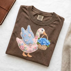 以下が含まれることがあります： 青い花のカゴを持ったガチョウのアップリケが特徴の茶色のTシャツ。ガチョウはピンクと青の花柄の翼と体を持っています。Tシャツはクルーネックと半袖です。Comfort Colorsブランドのラベルが見えます。