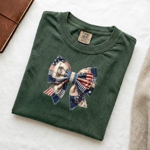 Puede incluir: Camiseta verde oscuro con un lazo patriótico aplicado. El lazo presenta un diseño de patchwork con patrones de la bandera estadounidense y la Estatua de la Libertad. La camiseta tiene cuello redondo y una etiqueta Comfort Colors.