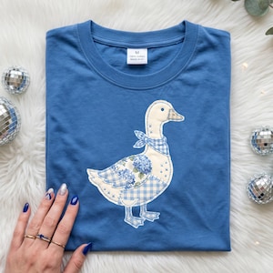 以下が含まれることがあります： 青いTシャツ。 遊び心のあるアヒルのアップリケが付いています。 アヒルは白い体、青いギンガムチェックのスカーフ、花の羽が特徴です。 シャツはクルーネックで、「M 100% Cotton MADE IN EU」と書かれたタグが付いています。