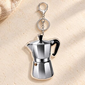 May include: A silver-toned acrylic keychain shaped like a classic moka pot coffee maker. The design features a metallic gray body with a black handle and a small gold-colored valve detail. It is attached to a silver metal split ring and lobster clasp.