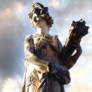 May include: A weathered stone statue of a woman holding a bundle of wheat. The sculpture is draped in classical robes and has a detailed hairstyle. The background features a cloudy sky with hints of sunlight.
