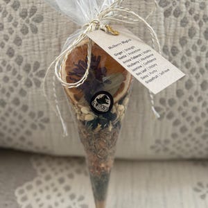 May include: A cone-shaped package filled with dried herbs and spices, tied with twine and a tag. The tag reads "Mulberry Magick" and lists ingredients like ginger, nettle, and jasmine. A dried orange slice is visible at the top. The package is clear.