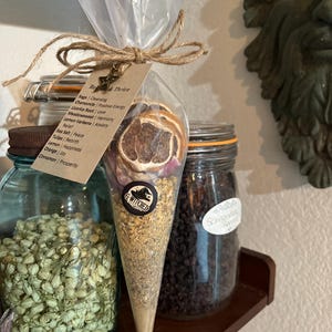 May include: A clear cellophane cone filled with dried herbs, flowers, and citrus slices, tied with twine and a tag that reads "Re&Thrive". A black and white sticker with the text "G-WITCHED" is on the cone. Jars of herbs and spices are in the background.