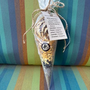 May include: A clear, cone-shaped package tied with twine, filled with dried herbs and botanicals. A tag lists ingredients like White Willow Bark, Jasmine, and Lavender. The package has the text "G-WITCHED" on it.