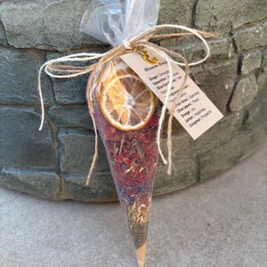 May include: A clear cone-shaped pouch filled with dried herbs, flowers, and a dried orange slice. The pouch is tied with twine and a tag that reads "Blossom Brews" and lists the ingredients and their associated benefits.
