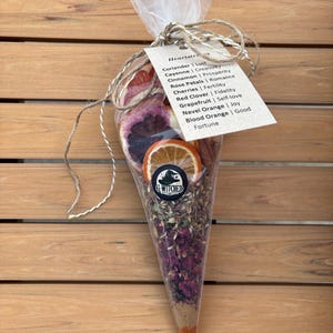 May include: A cone-shaped bag filled with dried fruit and herbs. The bag is tied with twine and has a label that reads "Heartstrings" and lists various herbs and their associated meanings. The bag is sitting on a wooden surface.