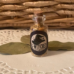 Puede incluir: Una pequeña botella de vidrio con un tapón de corcho llena de una sustancia marrón claro. La botella tiene una etiqueta en blanco y negro con un sombrero de bruja y el texto "G-Witched Earthy Remedies".