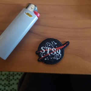 Puede incluir: Un encendedor BIC gris con un botón de encendido rojo y un parche negro de la NASA STS-9. El parche es circular, con texto blanco y una línea roja.
