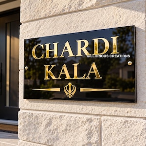 Könnte beinhalten: Ein rechteckiges schwarzes Schild mit goldener Schrift, die "CHARDI KALA" und "GILLORIOUS CREATIONS" lautet, sowie einem goldenen Khanda-Symbol unter dem Text. Das Schild ist mit goldenen Beschlägen an einer hellen Steinmauer befestigt.